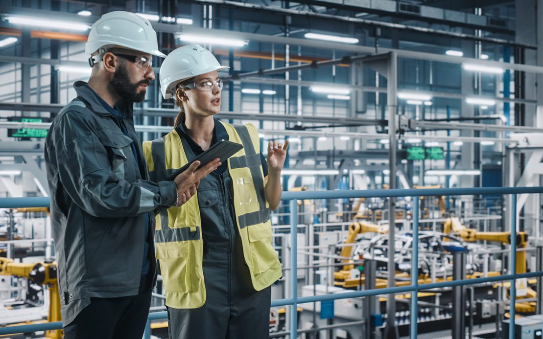Male Specialist and Female Car Factory Engineer in High Visibility Vests Using Tablet Computer. Automotive Industrial Manufacturing Facility Working on Vehicle Production. Diversity on Assembly Plant.