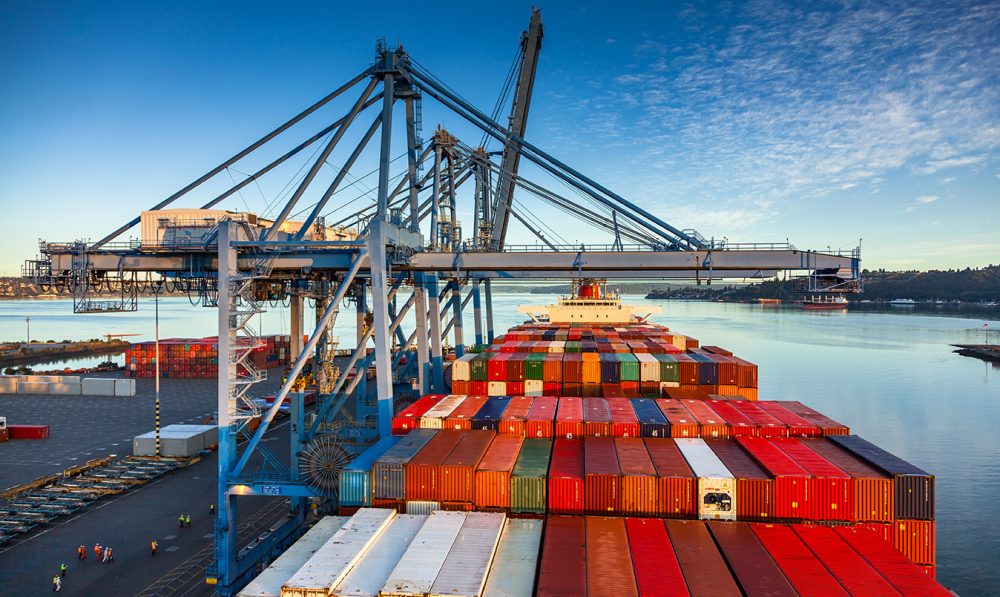 A high angle still image of a cargo ship with numerous containers on Blair Waterway, parked on container terminal in the Port of Tacoma, Washington on a sunny day.