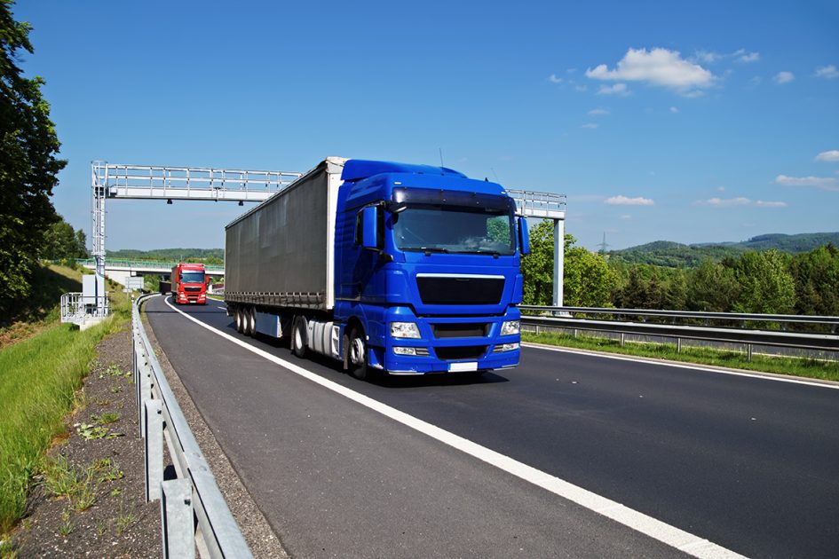 Blue truck passing through the electronic toll gates on the highway in a wooded landscape. Red truck, bridge and forested mountains in the background. White clouds in the blue sky.